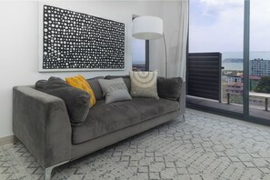 Ein graues Sofa mit Kissen, eine Wandkunst mit Muster, eine Stehlampe und ein Blick auf Gebäude durch ein Fenster.