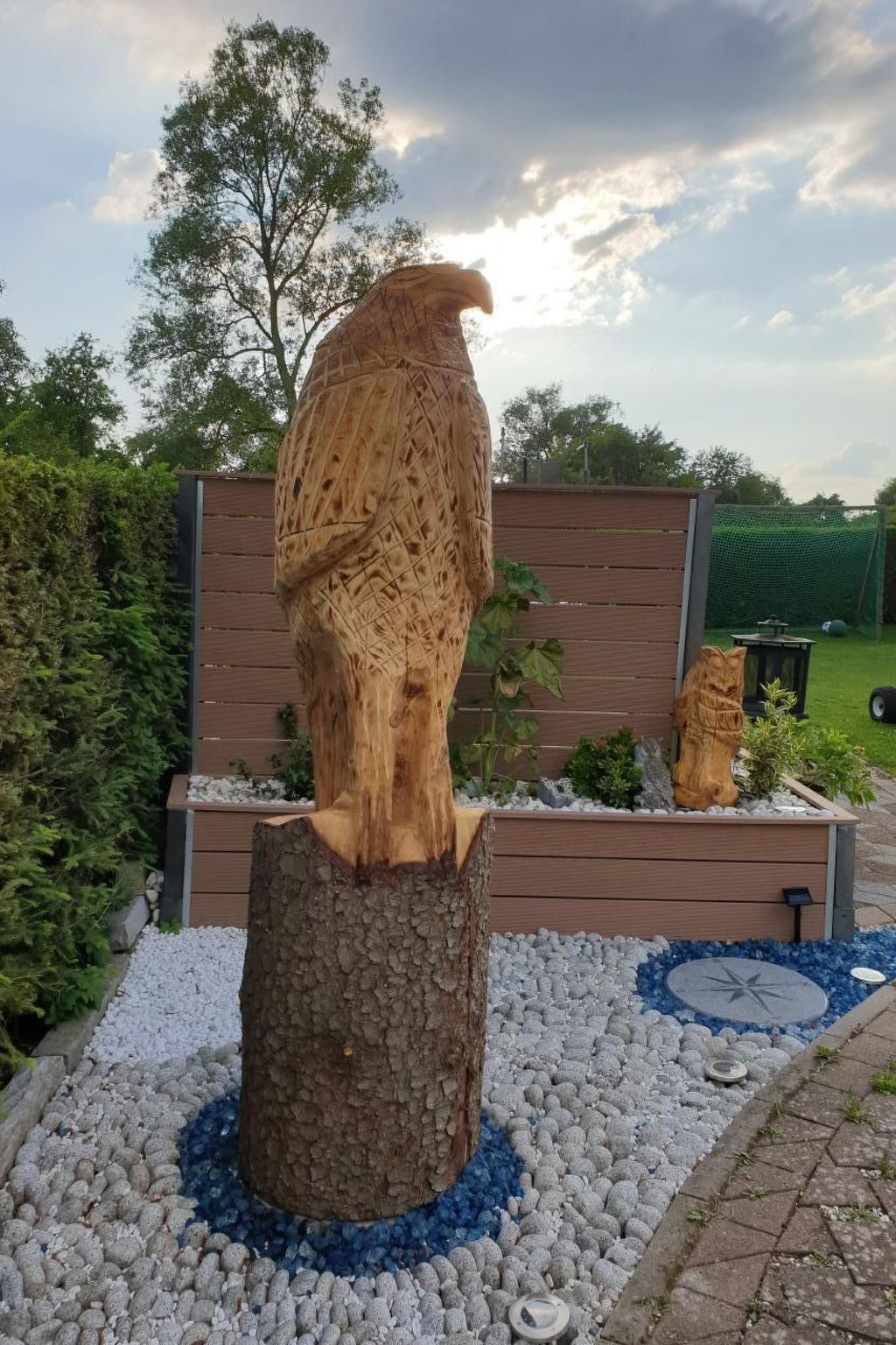 A wooden owl sculpture on a tree stump with a pebble border, set in a garden with a wooden fence and greenery in the background.