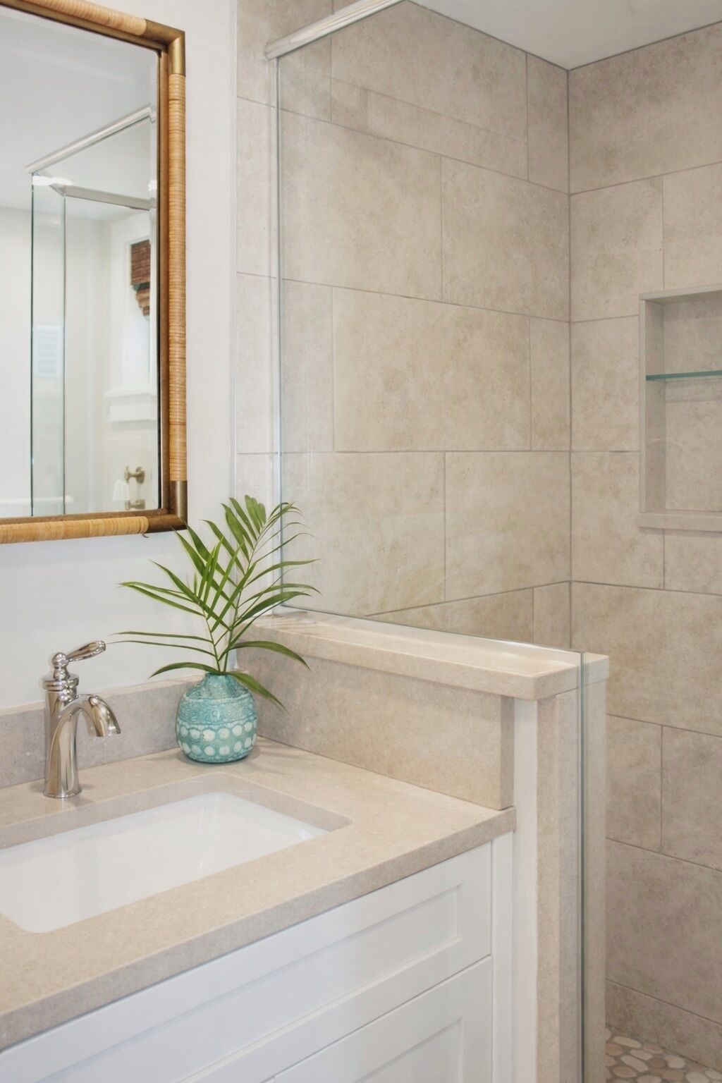 A bathroom with a beige tiled wall, a white sink with a silver faucet, a blue vase with a plant, and a mirror with a wooden frame.