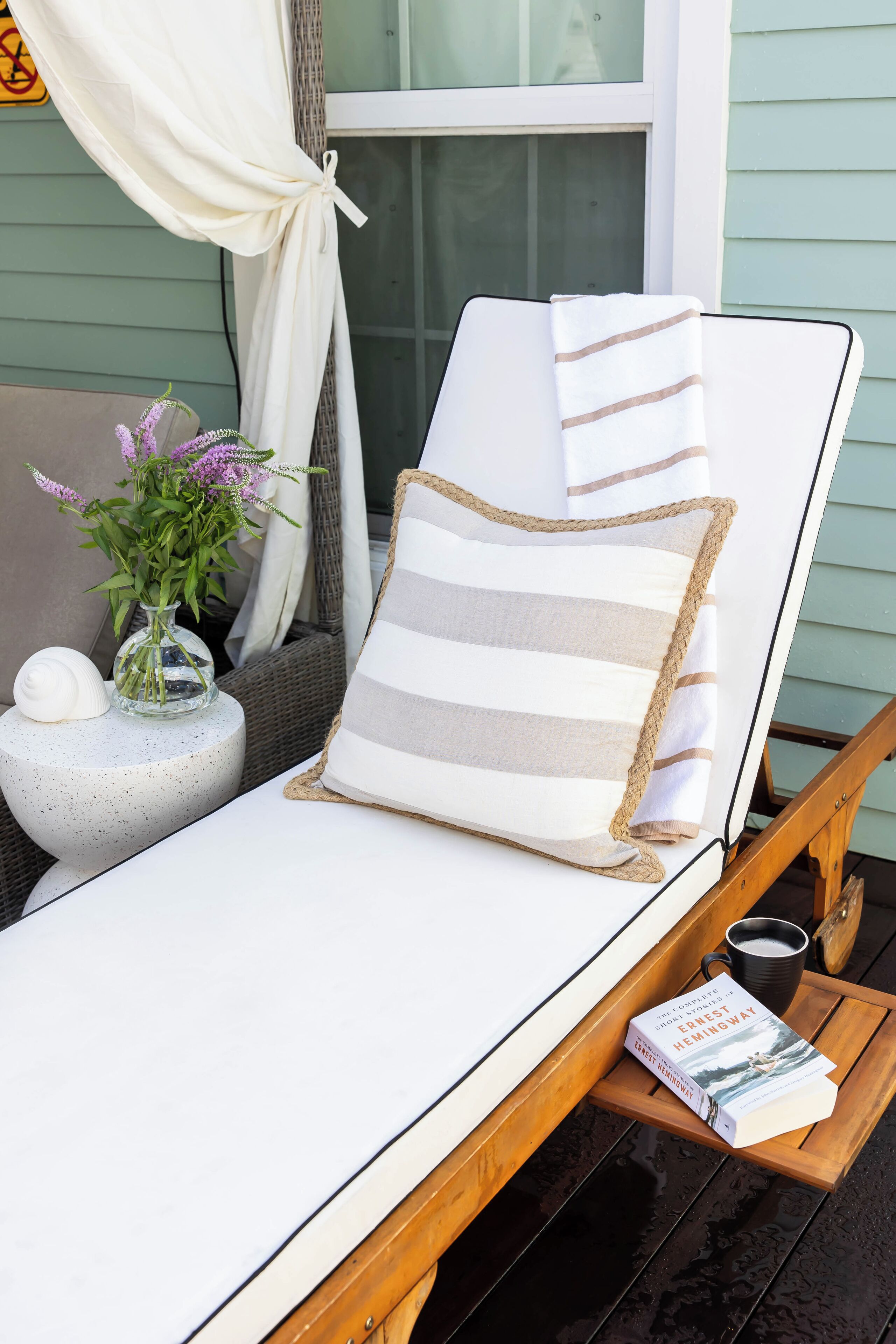 Un transat avec des coussins à rayures, un petit vase avec des fleurs et un livre posé sur un plateau en bois.