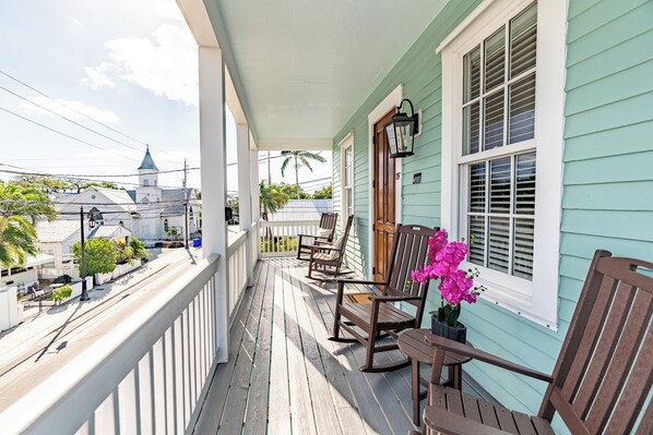 A porch with rocking chairs and a potted plant, overlooking a street with buildings and a church steeple. - Lighthouse Suites Collection (Key West)