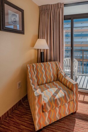 A hotel room with a patterned armchair, a lamp, a framed picture, and a view of a building through a window. - Landmark 1006 (Myrtle Beach)