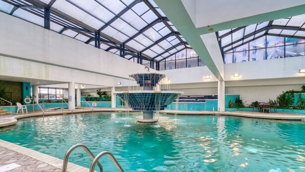 An indoor swimming pool with a central fountain, clear blue water, and a glass ceiling. - Landmark 1006 (Myrtle Beach)