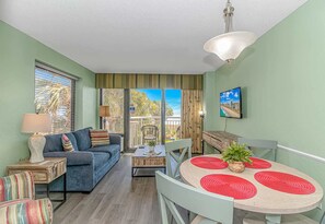 A room with a sofa, a dining table with red placemats, a TV on the wall, and a window with blinds. - Sailfish 210 (Myrtle Beach)
