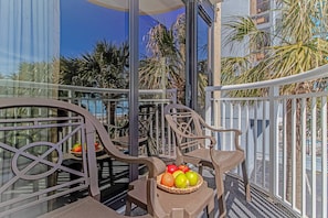 Ein Balkon mit einem Korbwäsche voller Obst, Metallstühlen und Blick auf Palmen und ein Gebäude.