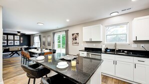 A modern kitchen with white cabinets, a black countertop, and a dining area with a table and chairs. - Shuffleboard~Hot Tub~Arcade~Foosball~Spacious for Groups~7 Min to Ruoff (Noblesville)