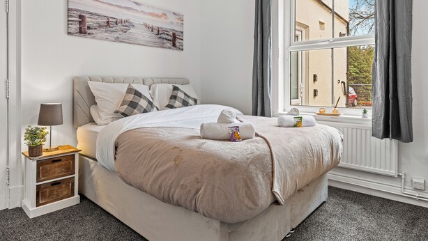 Una cama bien hecha con manta, almohadas y una toalla, una mesita de noche con una lámpara y una planta pequeña, una ventana con cortinas y un cuadro en la pared.