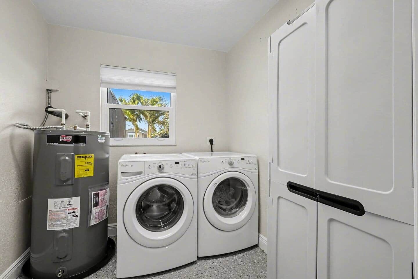 A compact laundry room with two front-loading washing machines, a water heater, and a window with a view of palm trees.