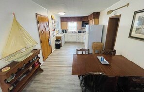 Une cuisine compacte avec des meubles blancs, un réfrigérateur et un four à micro-ondes. Une table à manger en bois, entourée de chaises, se trouve au premier plan.