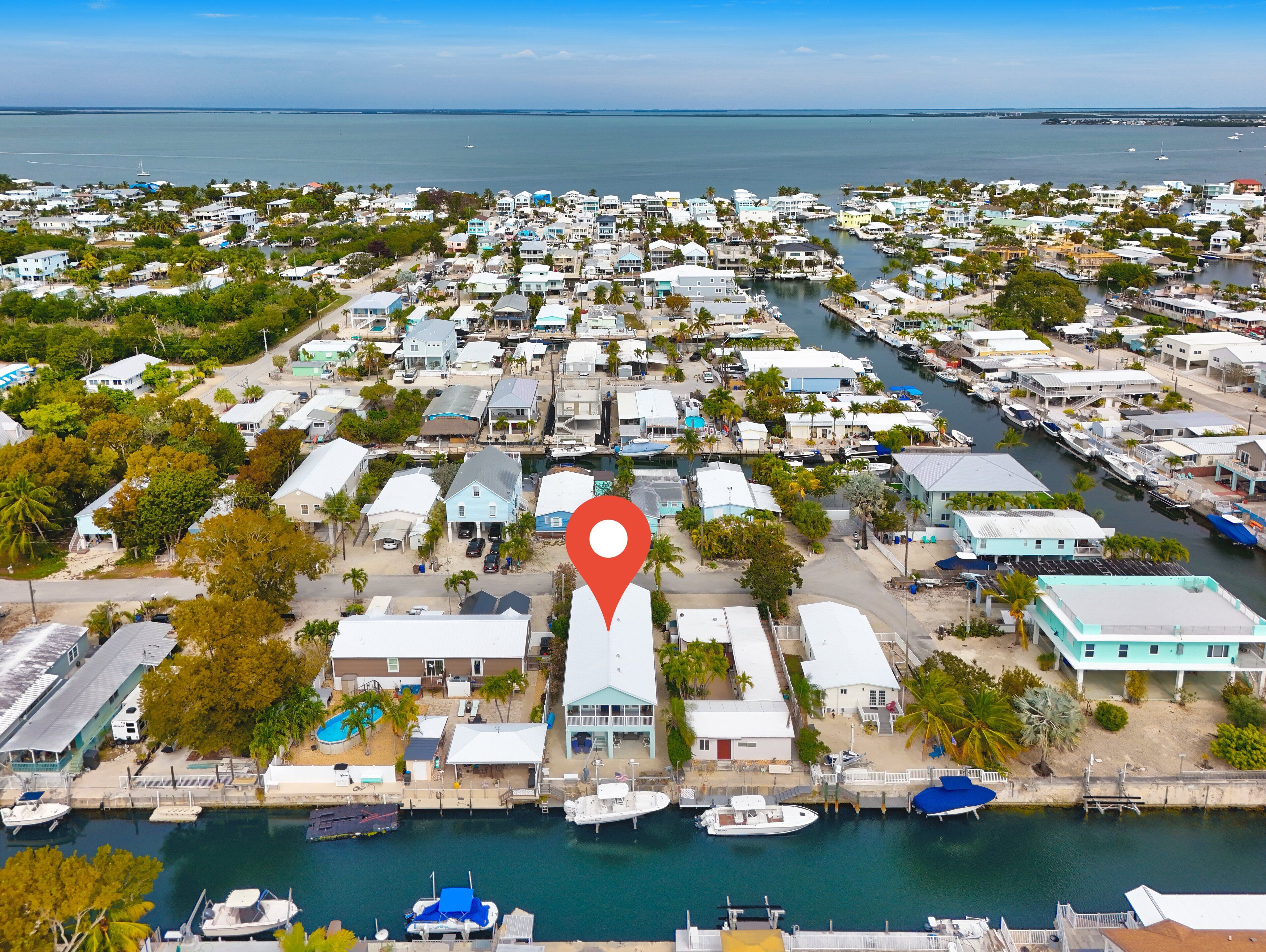 An aerial view of a coastal town with a marina, featuring numerous houses, boats, and a prominent red location marker.