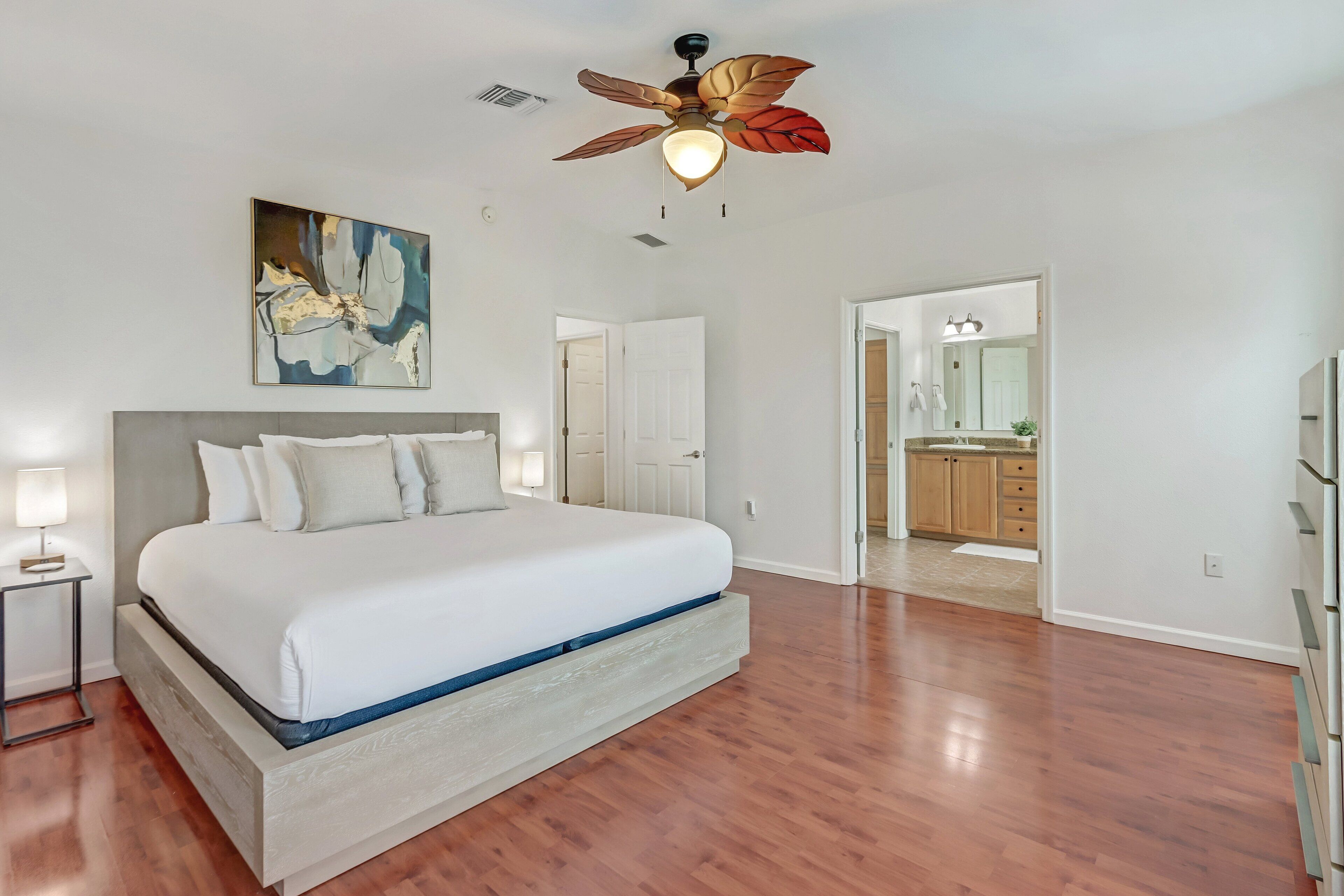 A bedroom with a large bed, a ceiling fan, a painting, and a view into the bathroom.