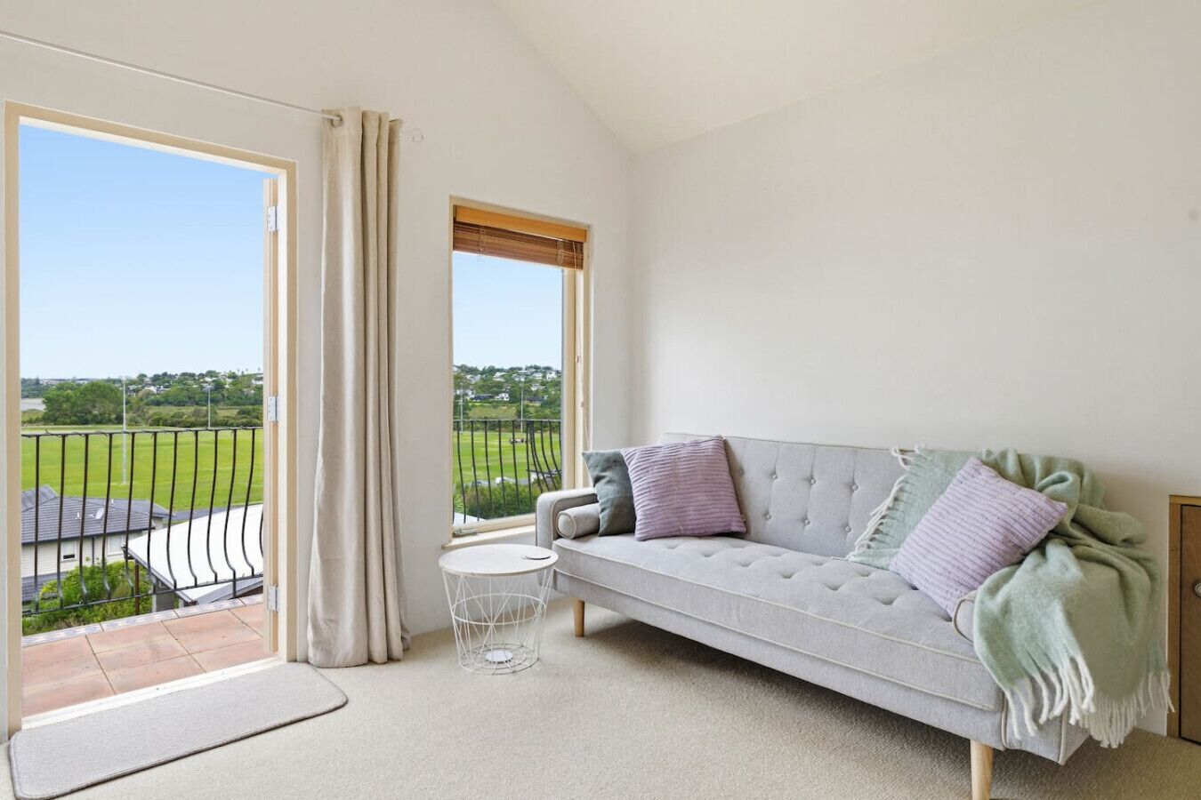 Ein geräumiges Wohnzimmer mit einem grauen Sofa, einem kleinen runden Tisch und einer großen Schiebeglasdoor zum Balkon mit Blick auf ein grünes Feld.