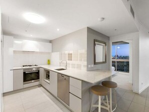 A modern kitchen with a central island, stainless steel appliances, and a view of the city through a large window. - Cozy 2BR Mosaic Valley | Views, Rooftop & Parking (Brisbane)