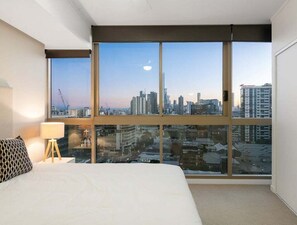 A hotel room with a large window offering a city view, a bed, a bedside lamp, and a patterned pillow. - Cozy 2BR Mosaic Valley | Views, Rooftop & Parking (Brisbane)