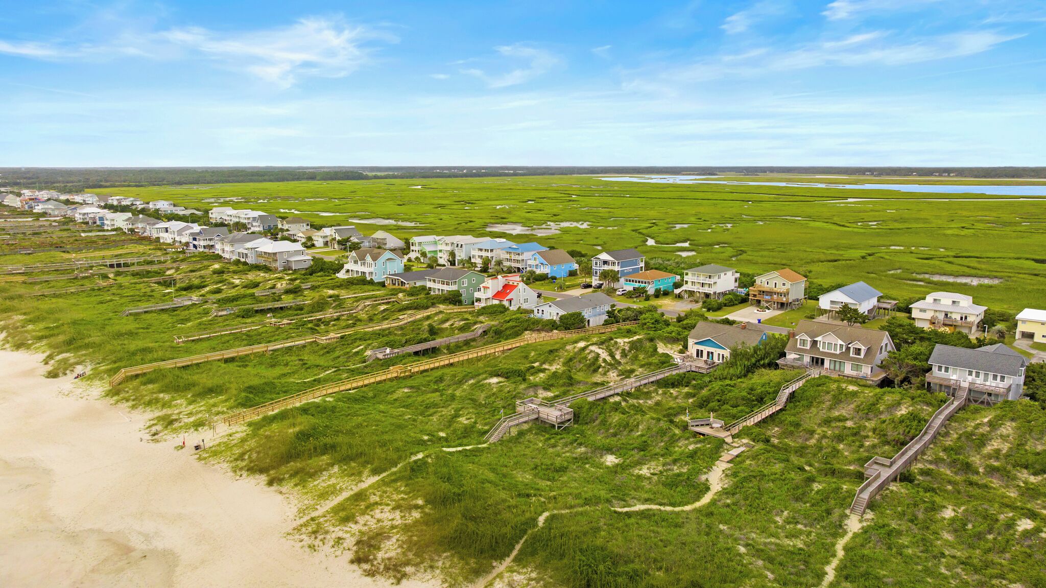 Vista aérea de una zona residencial costera con casas conectadas por pasarelas de madera, una playa y un paisaje pantanoso.