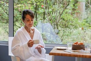 A person in a white bathrobe sitting by a window with a table that has a croissant basket and a cup. - La rochebardière apartment (SAINT PAUL LES DAX)