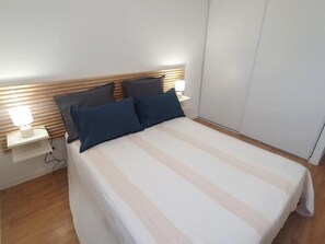 A neatly made bed with a striped bedspread, two pillows, and a bedside lamp on a white nightstand. - La rochebardière apartment (SAINT PAUL LES DAX)