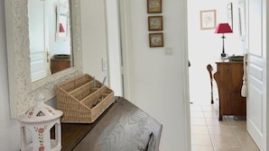 Um corredor com cômoda de madeira, espelho e cesta de vime. Quadros emoldurados na parede e um abajur vermelho ao fundo.