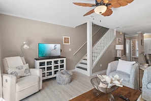 A living room with a sofa, ottoman, TV, cabinet, and a ceiling fan. - Seacrest West - 7 Palms 3 (Panama City Beach)