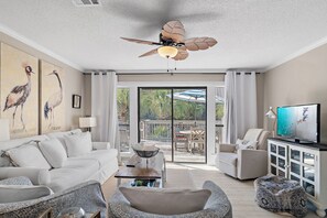 Un soggiorno con ventilatore a soffitto, un divano, un tavolino da caffè, una TV e un balcone con mobili da esterno.