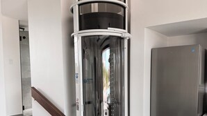 A modern kitchen with a built-in stainless steel refrigerator, a curved glass elevator shaft, and a staircase with grey tiles. - Luxury Bungalow + Private Infinity Pool + Firepit (Long Bay Hills)