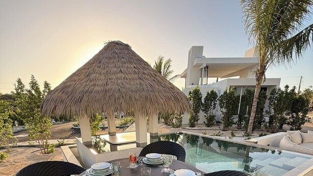A round table set for dining outdoors with a thatched-roof structure in the background, a pool, and modern buildings. - Luxury Bungalow + Private Infinity Pool + Firepit (Long Bay Hills)