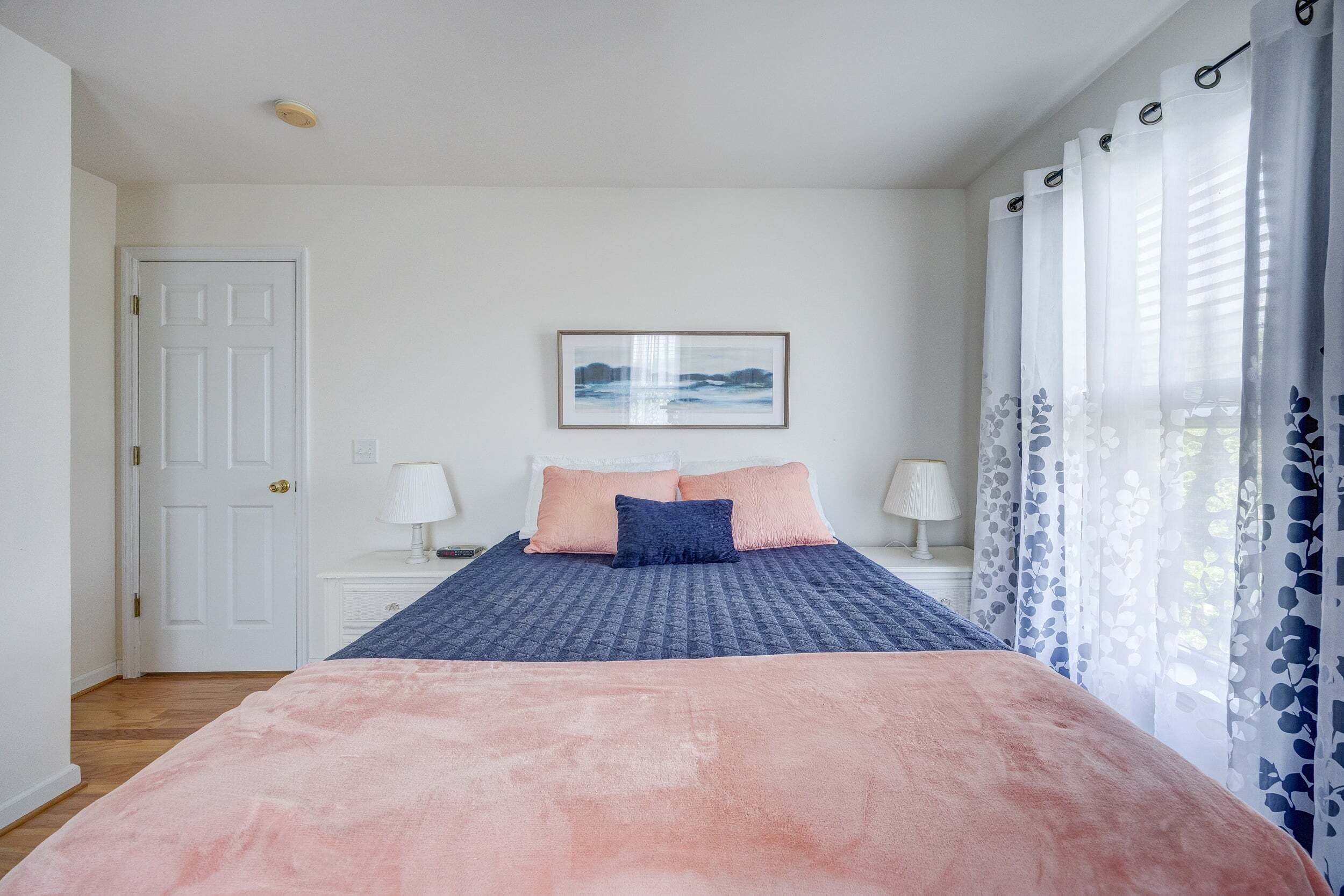 A bedroom with a bed, a door, a nightstand with a lamp, and a framed picture on the wall.
