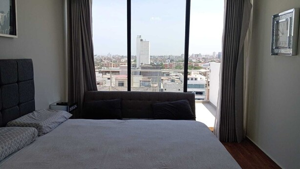 Un dormitorio con una cama grande, cabecera, mesita de noche y vistas de la ciudad a través de ventanas amplias.