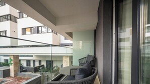 Ein Balkon mit Korbstuhl, Kissen, Glasgeländer und Blick auf benachbarte Gebäude.