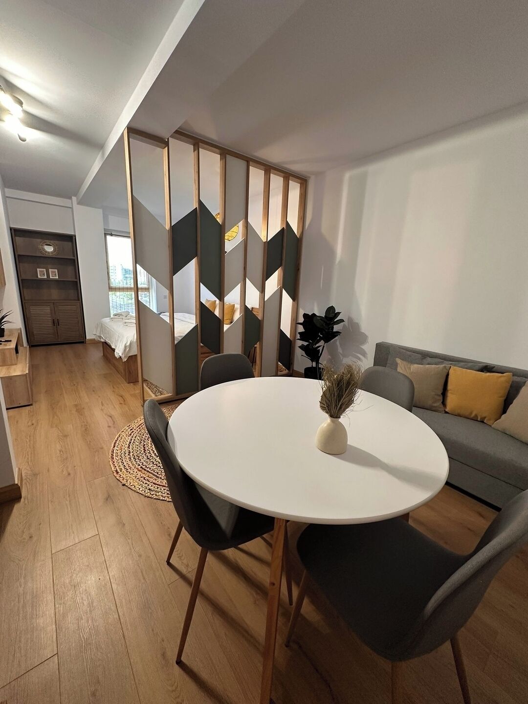 A modern living room with a round white table, two black chairs, a grey sofa with yellow cushions, and a wooden floor.