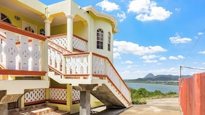 A multi-story building with a staircase leading to a balcony, a view of a body of water, and a mountainous landscape in the distance. - 2BR Oceanview • Walk to Beach • Saint Lucia (Vieux Fort)