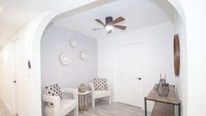 Une pièce avec une porte blanche, un ventilateur de plafond et deux chaises blanches dotées de coussins à motifs.