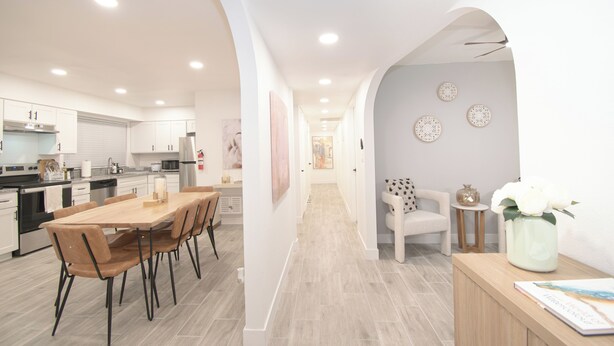 Une cuisine moderne avec des meubles blancs, un coin repas avec une table et des chaises en bois, et un couloir agrémenté d’un canapé blanc et d’assiettes décoratives accrochées au mur.