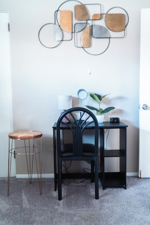 Ein kleiner Arbeitsraum mit Schreibtisch, Stuhl und Hocker. Darüber hängt ein abstraktes Kunstwerk an der Wand.