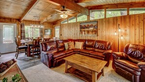 A wooden cabin interior with a large leather sectional sofa, a wooden coffee table, and a dining area with a table and chairs.