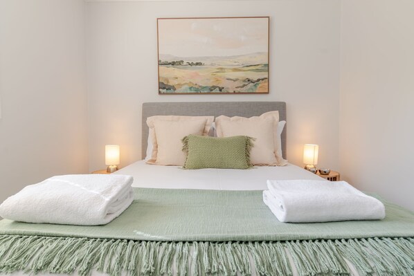 Un lit bien fait, avec du linge de lit blanc, un plaid vert et deux serviettes blanches. Au-dessus du lit, on peut voir un tableau représentant un paysage, encadré, et de chaque côté du lit, il y a deux lampes de chevet.