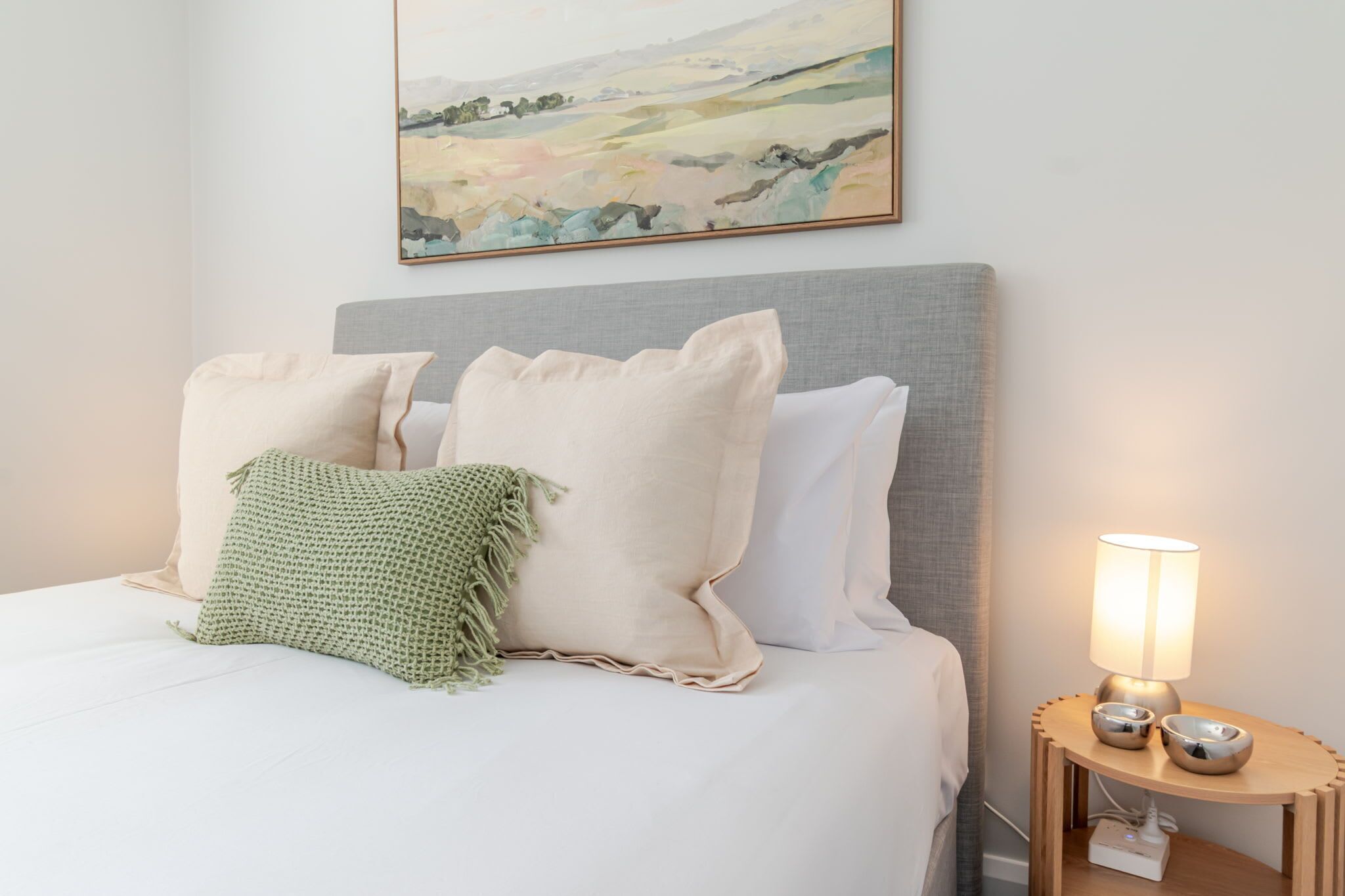 Un lit bien fait, avec du linge de lit blanc et plusieurs oreillers, une lampe de chevet et une petite table de nuit en bois sur laquelle reposent deux bols.