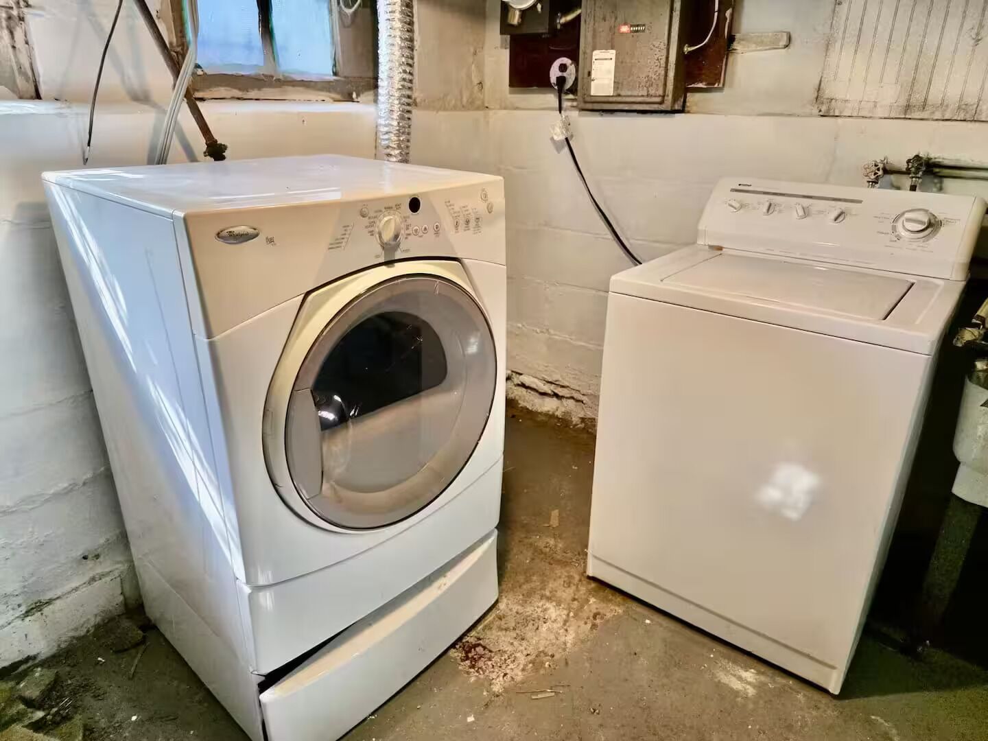 Une buanderie avec deux machines à laver blanches et un sèche-linge, dotée de murs en béton apparent et d’une fenêtre.