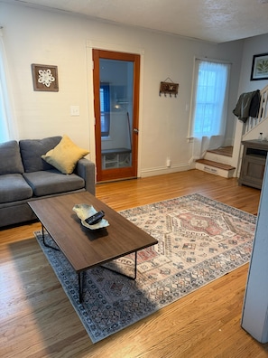 Una sala de estar con un sofá gris, una mesa de centro de madera, un tapete estampado y una escalera que conduce a otra habitación.