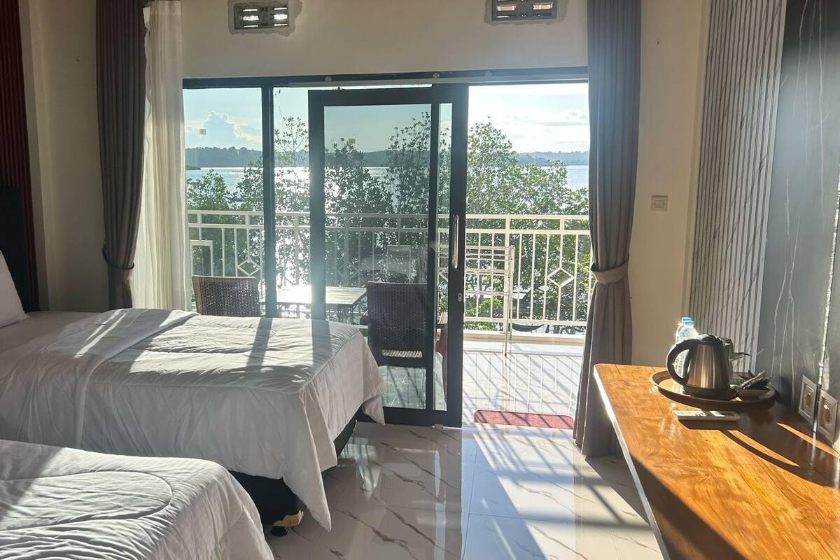A hotel room with two beds, a balcony with a view of the sea, and a wooden table with a kettle.