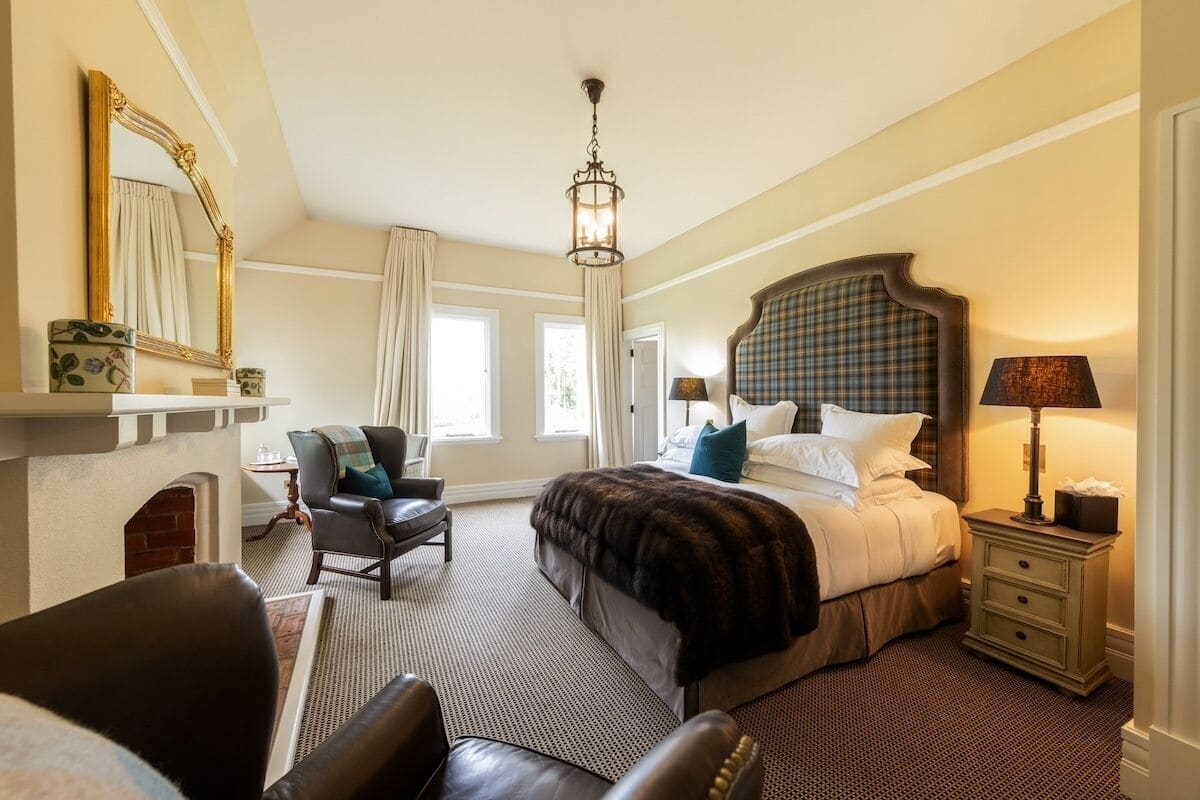 A hotel room with a large bed, a chair, a nightstand, a lamp, a fireplace, and a mirror.