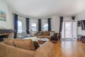 Een woonkamer met een bank, fauteuil en een salontafel. Er is een open haard, een televisie en een deur naar buiten.