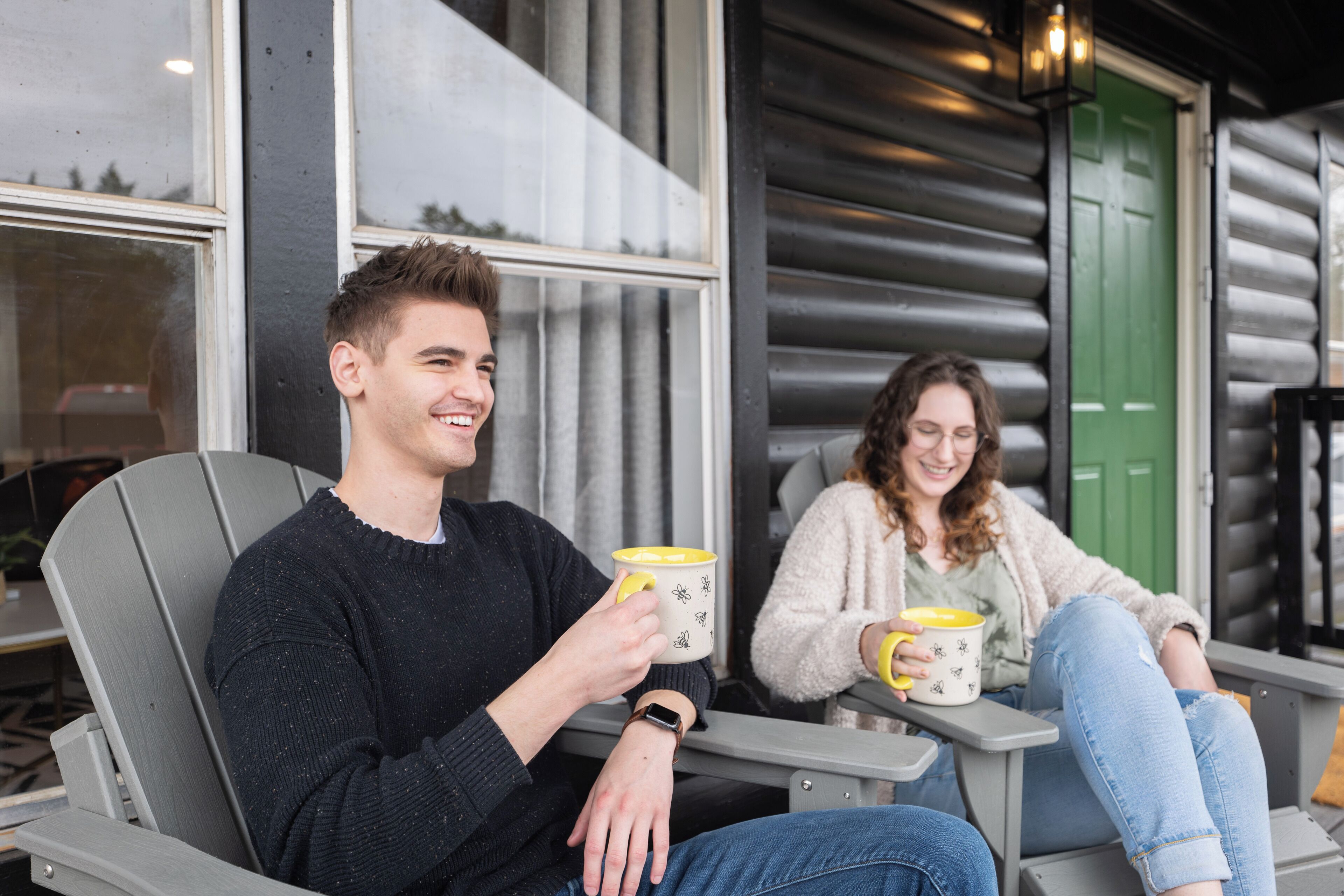 Two people sitting on outdoor chairs, holding mugs, with a log cabin in the background.