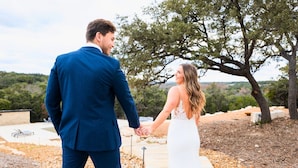Una pareja tomados de la mano en un camino de piedra, con un árbol y un edificio al fondo.