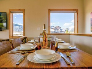 Une table à manger dressée avec des assiettes, des verres, des couverts et une bouteille, et des fenêtres donnant sur une vue sur les montagnes.