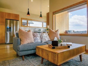 A modern kitchen with a stainless steel refrigerator, a gray sofa with decorative pillows, a wooden coffee table, and a large window offering a view of the sky and clouds. - Scandinavian Condo w/ Mountain Views & Free Parking (Crested Butte)