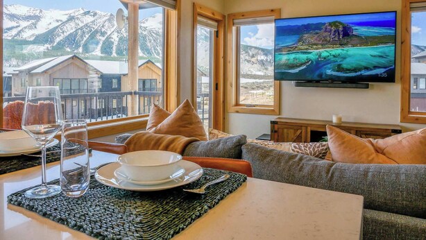 Habitació amb una finestra gran que ofereix vistes a la muntanya, televisió de pantalla plana i zona de menjador amb taula preparada per a dues persones.