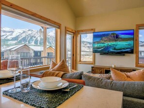 Ein Zimmer mit großem Fenster und Blick auf die Berge, einem Flachbildfernseher und einer Essecke mit Tisch für zwei Personen.