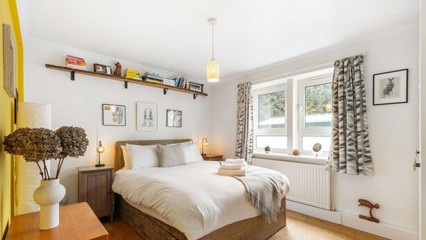 Ein Schlafzimmer mit einem hölzernen Bett, Nachttischen, einem Fenster mit Vorhängen, einem Regal mit Büchern und gerahmten Bildern an der Wand.