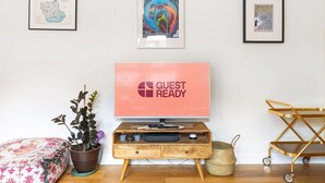 Ein Zimmer mit Fernseher, der 'GUEST READY' anzeigt, einem Holztischchen, einer Pflanze, einem farbenfrohen Kissen und gerahmten Bildern an der Wand.